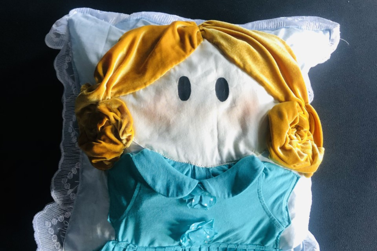 Zdjęcie przedstawia białą, kwadratową poduszkę na czarnym tle. Na poduszkę naszyto prosty kształt popiersia małej dziewczynki w niebieskiej bluzce i żółtych włosach związanych w dwa koczki po bokach twarzy.