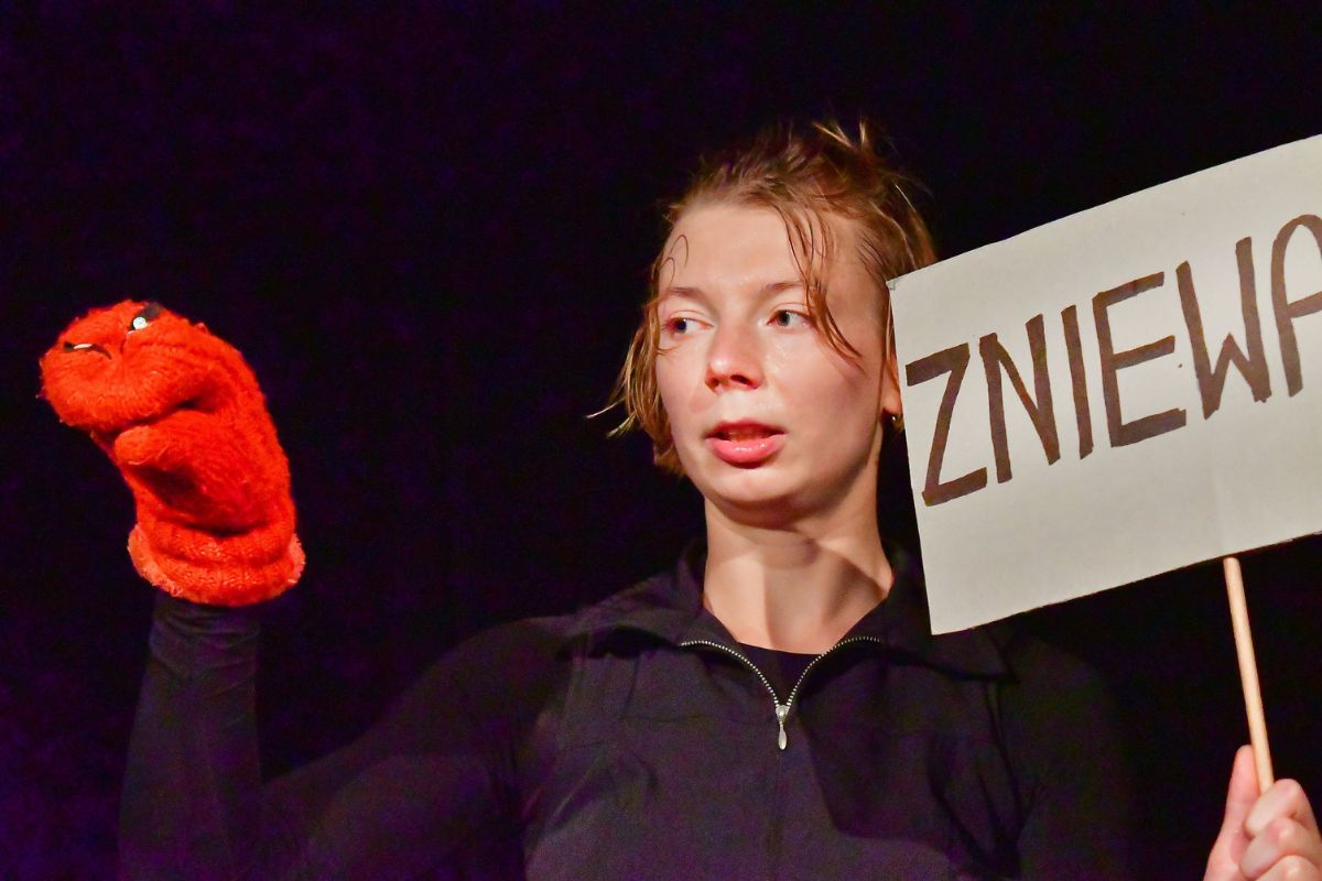 Zdjęcie przedstawia młodą aktorkę w bliskim planie ubraną na czarno na czarnym tle. Na prawej ręce ma pacynkę zrobioną z czerwonej rękawicy, w lewej trzyma transparent, na którym widać tylko fragment słowa o treści "zniewa"