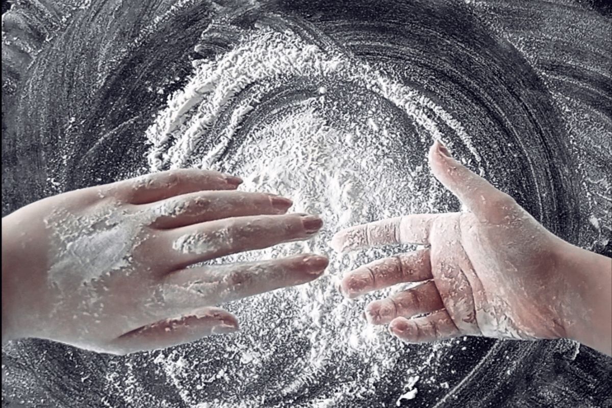 Zdjęcie przedstawia dłonie oprószoną mąką widoczne na tle czarnego stołu z mąką ułożoną w zamaszysty okrąg. Jedna z dłoni ułożona jest wierzchem do widza, druga wewnętrzną stroną.