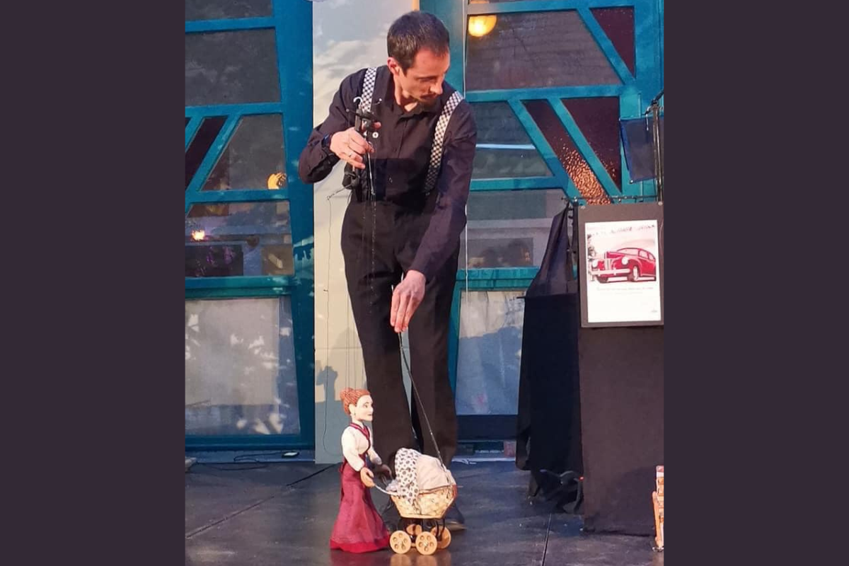Na zdjęciu Bence Sarkadi ubrany w czarną koszulę i czarne spodnie na szelkach animuje marionetką kobiety pchającej dziecięcy wózek. Mężczyzna stoi na scenie.