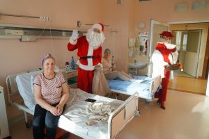 W szpitalnej sali dwie pacjentki siedzą na łóżkach, obok nich Święty Mikołaj z workiem na prezenty i elegancka Pani Mikołajowa w czerwonym kapeluszu z woalką.
