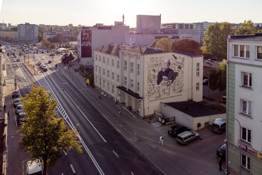 Widok muralu z oddalenia wraz z fragmentem ulicy i innych budynków.