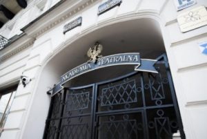 Zdjęcie fragmentu kutej bramy z pozłacanym napisem Akademia Teatralna oraz godłem, brama na froncie jasnego, zabytkowego budynku.