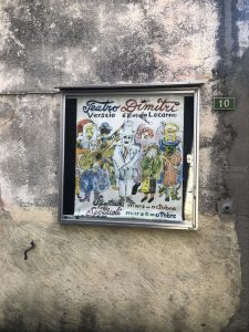 Zdjęcie umieszczonego w gablocie przyczepionej do muru plakatu promującego spektakle w Accademii Dimitri przedstawiającego lalki teatralne namalowane w technice akwareli.