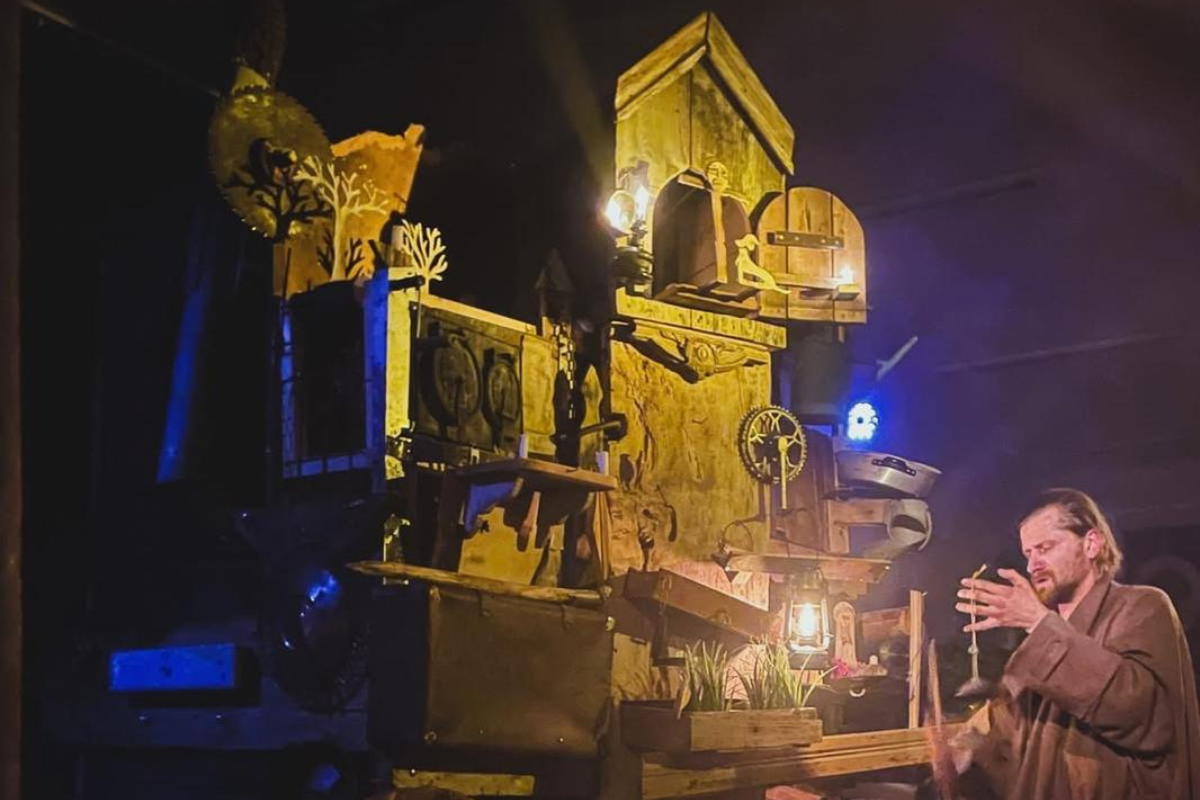 W prawym dolnym rogu młody mężczyzna w płaszczu. Obok niego drewniana konstrukcja scenograficzna w kształcie domu na kółkach.