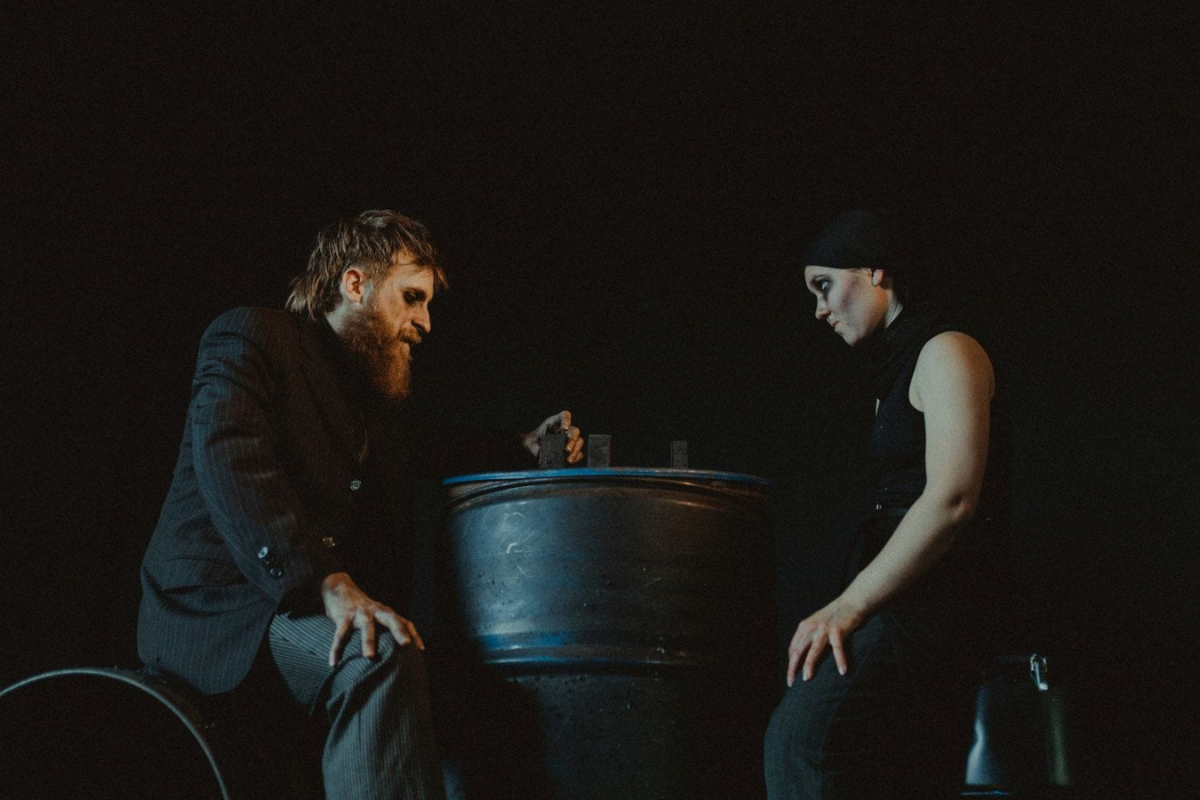 Na zdjęciu scena ze spektaklu: aktorzy - Daria Holovchanska i Marcin Tomkiel siedzą naprzeciwko siebie w kostiumach. Między nimi znajduje się duża metalowa miska.