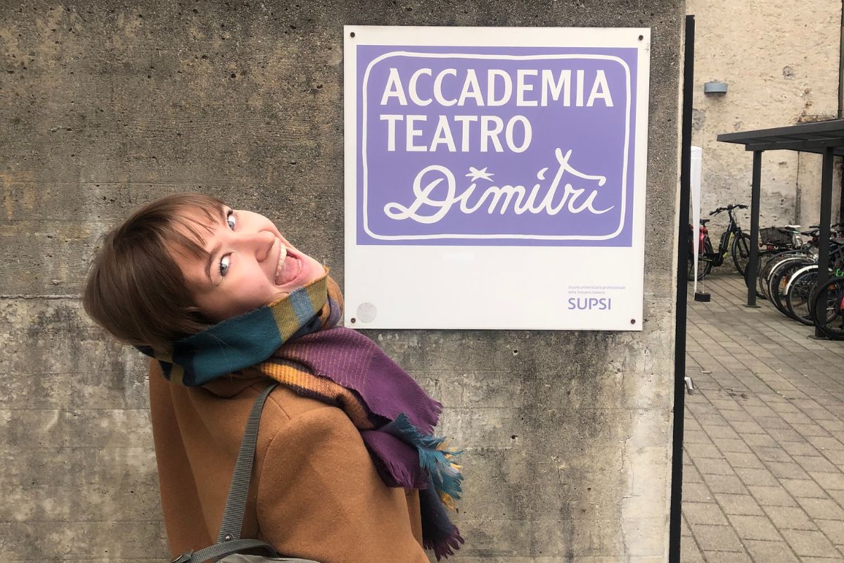 Studentka Karolina Klemczak stoi uśmiechnięta przy tablicy na murze z napisem "Accademia Teatro Dimitri".