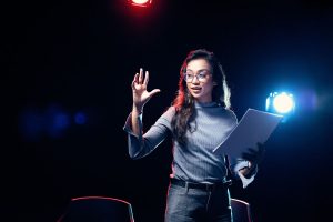 Młoda kobieta stoi na scenie z jedną ręką podniesioną do góry; w drugiej trzyma scenariusz.