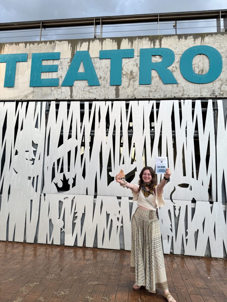 Zuzanna Praczuk stoi przed ażurową elewacją teatru i trzyma w rękach bilety oraz ulotkę spektaklu. 