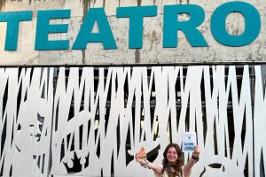 Zuzanna Praczuk stoi przed ażurową elewacją teatru i trzyma w rękach bilety oraz ulotkę spektaklu.