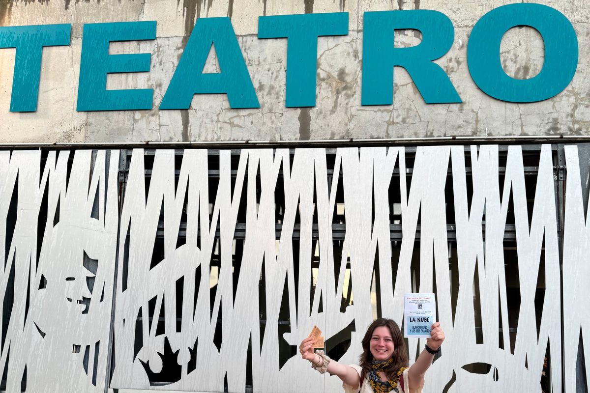 Zuzanna Praczuk stoi przed ażurową elewacją teatru i trzyma w rękach bilety oraz ulotkę spektaklu.