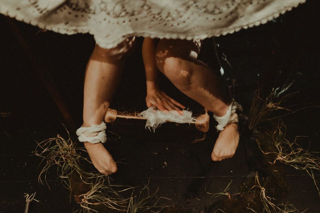 Dwie kobiece łydki sfotografowane od góry, kostki obwiązane białą tkaniną. Między łydkami kobieca ręka trzymająca skrawek koronkowego obrusa.