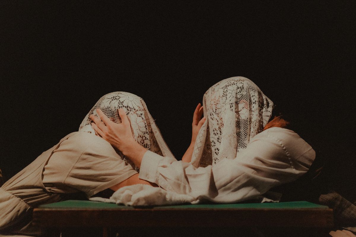 Dwie osoby siedzą naprzeciwko siebie przy stole, dotykając wzajemne swoich głów zakrytych koronkowym obrusem.