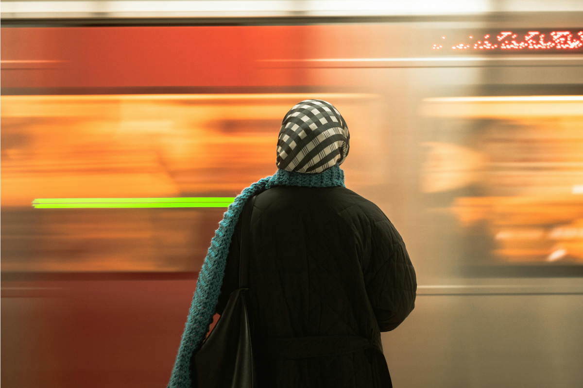 Zdjęcie kobiety w chustce na głowie widzianej od tyłu stojącej na peronie i obserwującej rozmyty przejeżdżający pociąg.