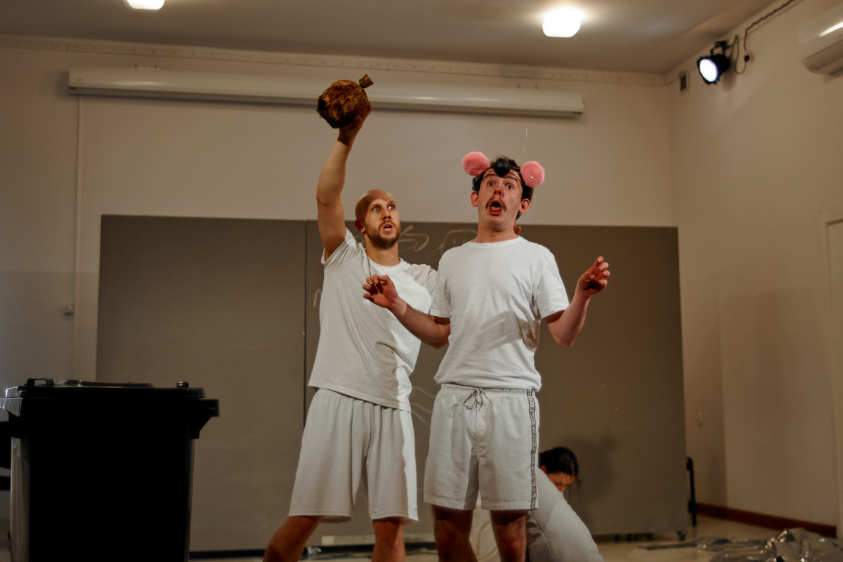 Dwóch młodych aktorów stoi na scenie w białych T-shirtach i krótkich spodenkach. Jeden trzyma w ręku element kostiumu niedźwiedzia i ma straszną minę, drugi - różowe uszy gryzonia i przerażoną minę.