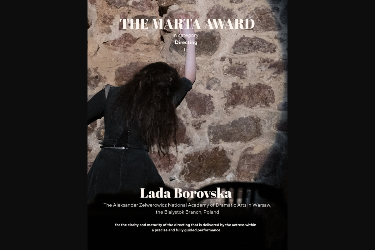 Zdjęcie z występu: postać młodej kobiety w czarnej sukni widziana od tyłu na tle kamiennej ściany.