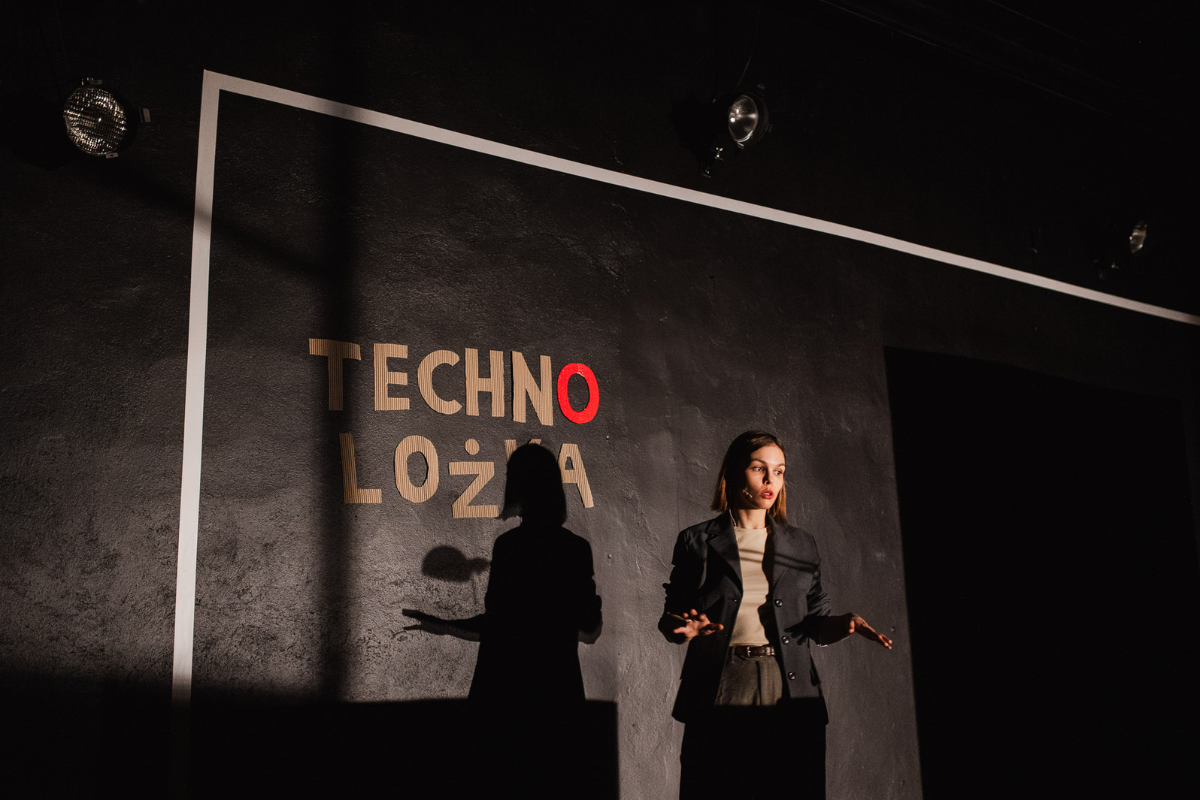 Młoda kobieta na scenie, za nią na ścianie tekturowy napis "technolożka".